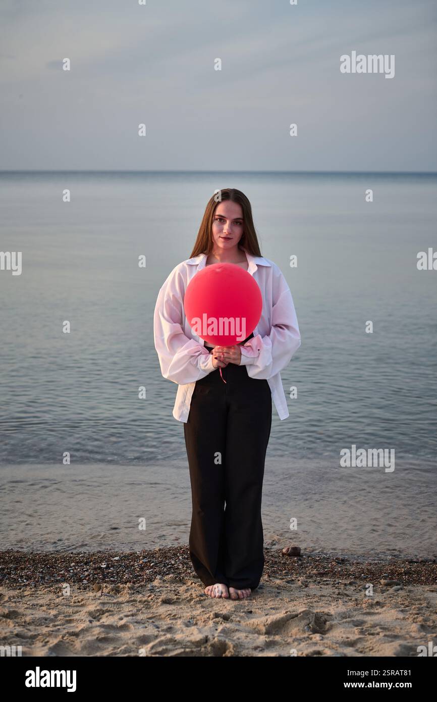 jeune femme se tient sur la plage sereine, tenant un ballon rouge vif. Mer calme et crépuscule doux créent une ambiance paisible et contemplative, symbolisant le solit Banque D'Images