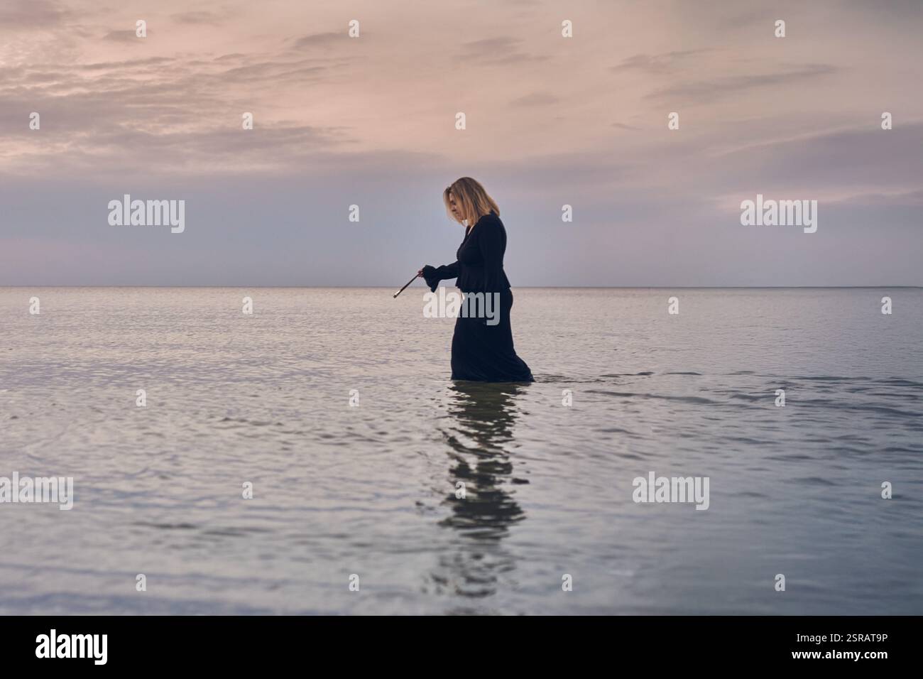 Jeune femme en tenue noire marche à travers les eaux calmes de l'océan au crépuscule, incarnant la solitude et la réflexion. Son comportement serein fait écho à l'immobilité de la mer Banque D'Images