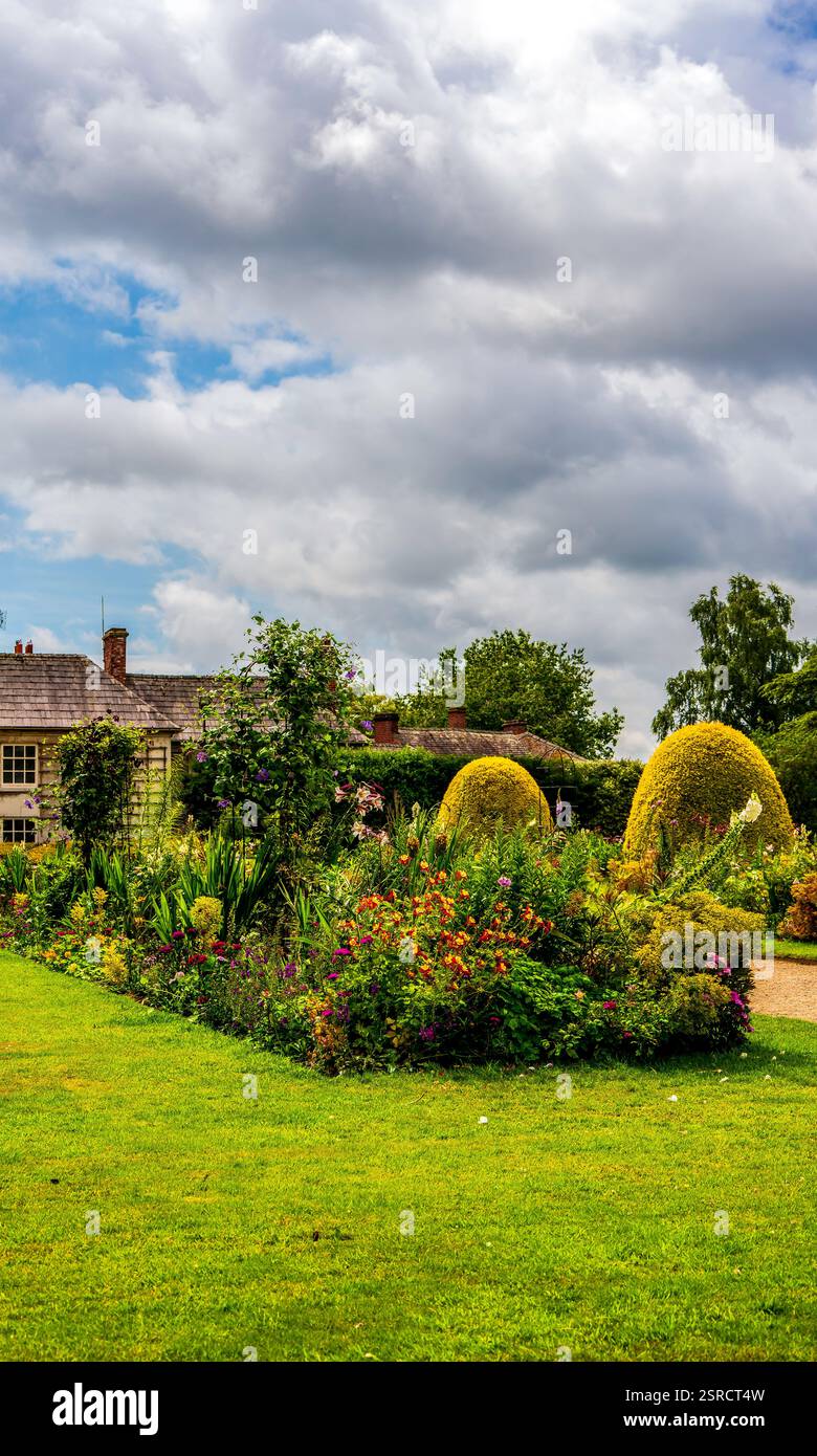 Jardin idyllique de Cottage anglais avec lits de fleurs vibrants et haies sculptées sous un ciel nuageux spectaculaire Banque D'Images