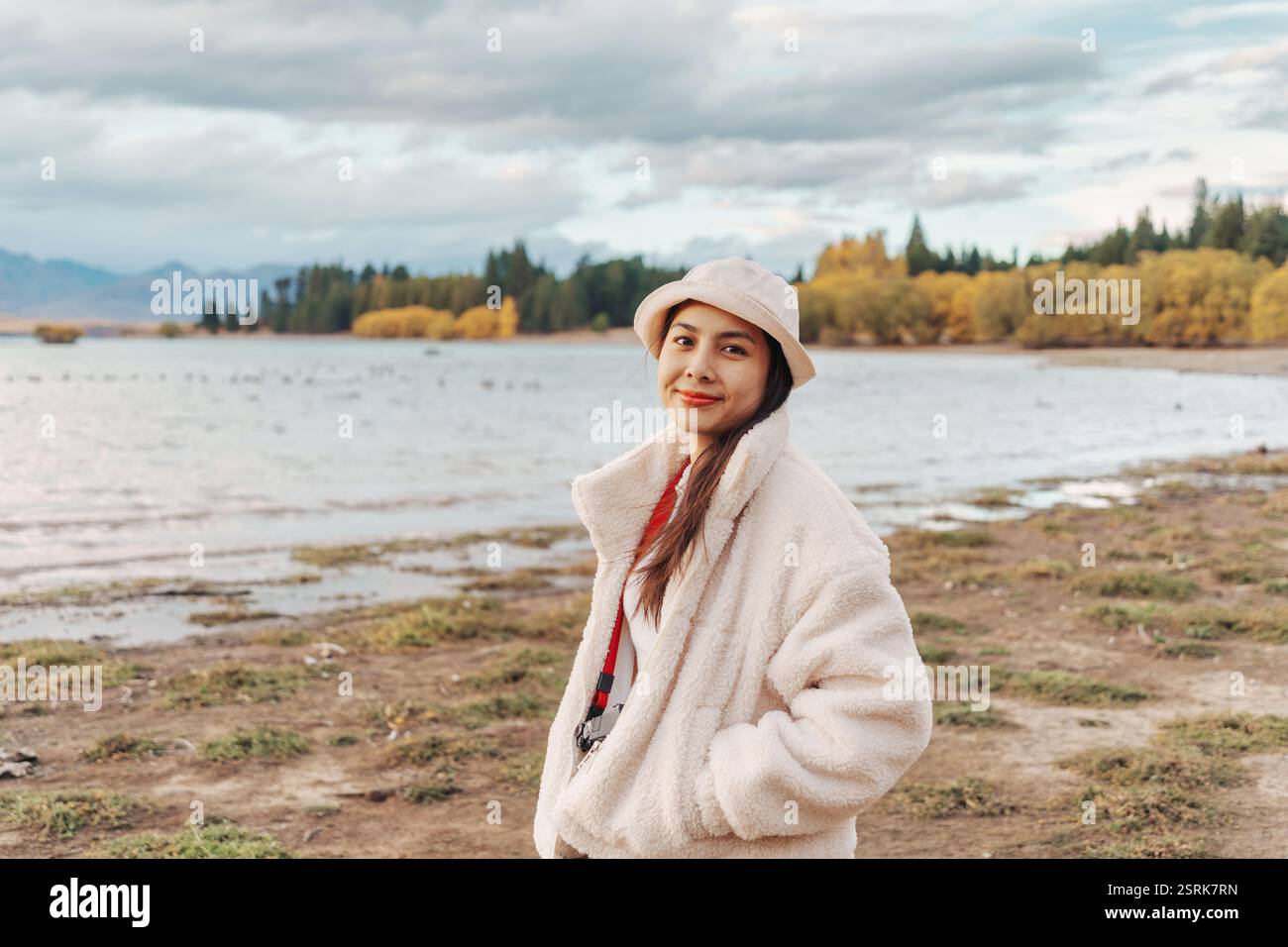 Jeune femme asiatique en veste confortable avec chapeau souriant et appréciant dans le parc d'automne par le lac Tekapo en Nouvelle-Zélande Banque D'Images