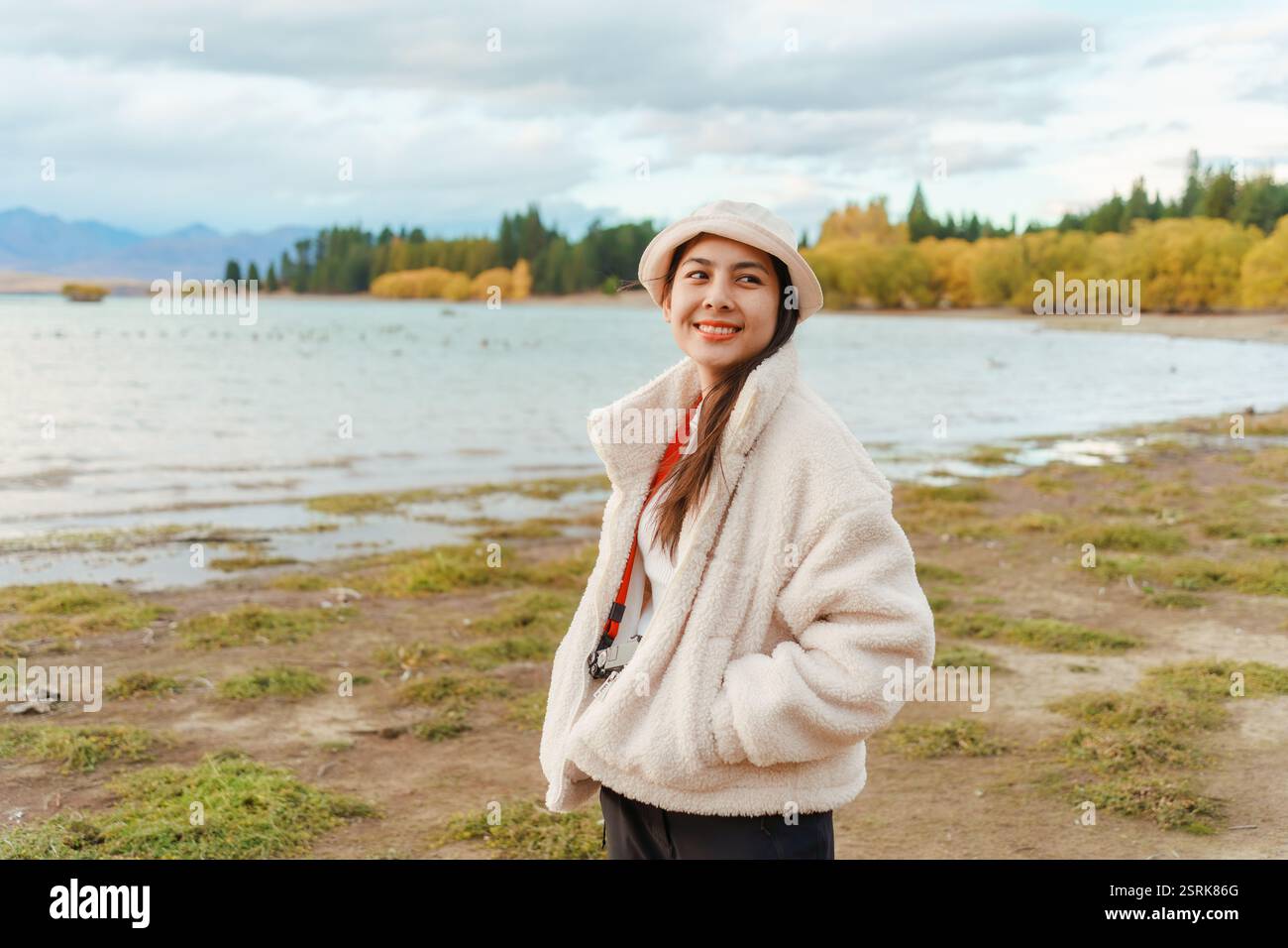 Jeune femme asiatique en veste confortable avec chapeau souriant et appréciant dans le parc d'automne par le lac Tekapo en Nouvelle-Zélande Banque D'Images