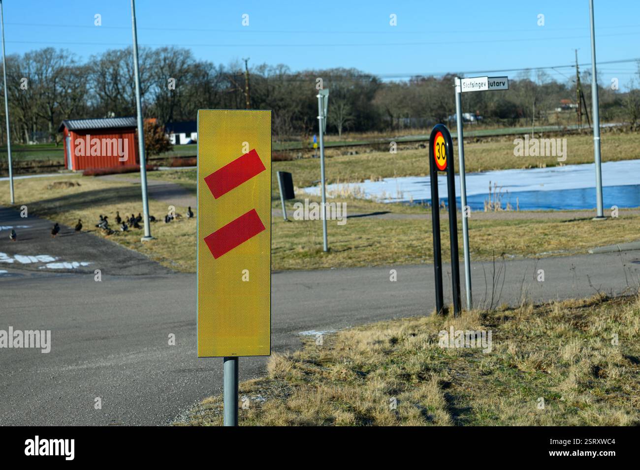 Un panneau routier jaune vif avec des chevrons rouges avertit les conducteurs qui ferment le chemin de fer. En arrière-plan, un bâtiment rouge et des canards peuvent être vus dans un rur paisible Banque D'Images Un panneau routier jaune vif avec des chevrons rouges avertit les conducteurs qui ferment le chemin de fer. En arrière-plan, un bâtiment rouge et des canards peuvent être vus dans un rur paisible Banque D'Images