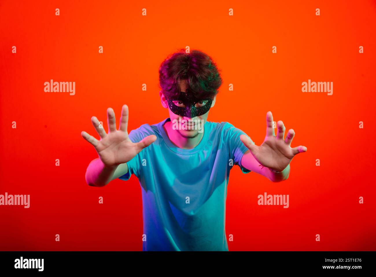 Heureux beau jeune homme portant un masque de carnaval regardant et faisant des gestes de la main à la caméra. Isolé sur fond de néon orange. Brésil Banque D'Images