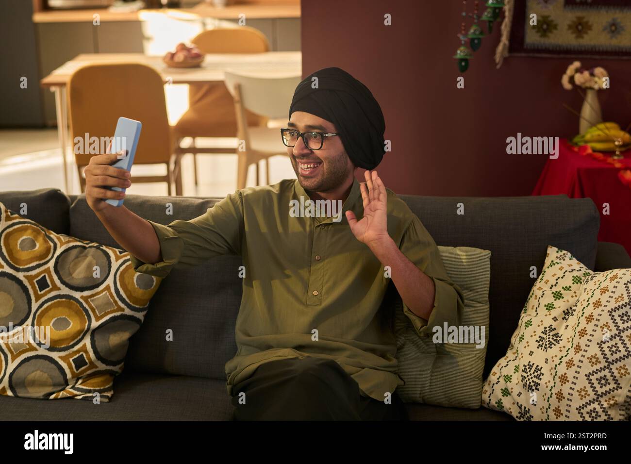 Homme assis sur le canapé tenant le smartphone et agitant tout en faisant un appel vidéo. Le décor moderne du salon est visible en arrière-plan Banque D'Images