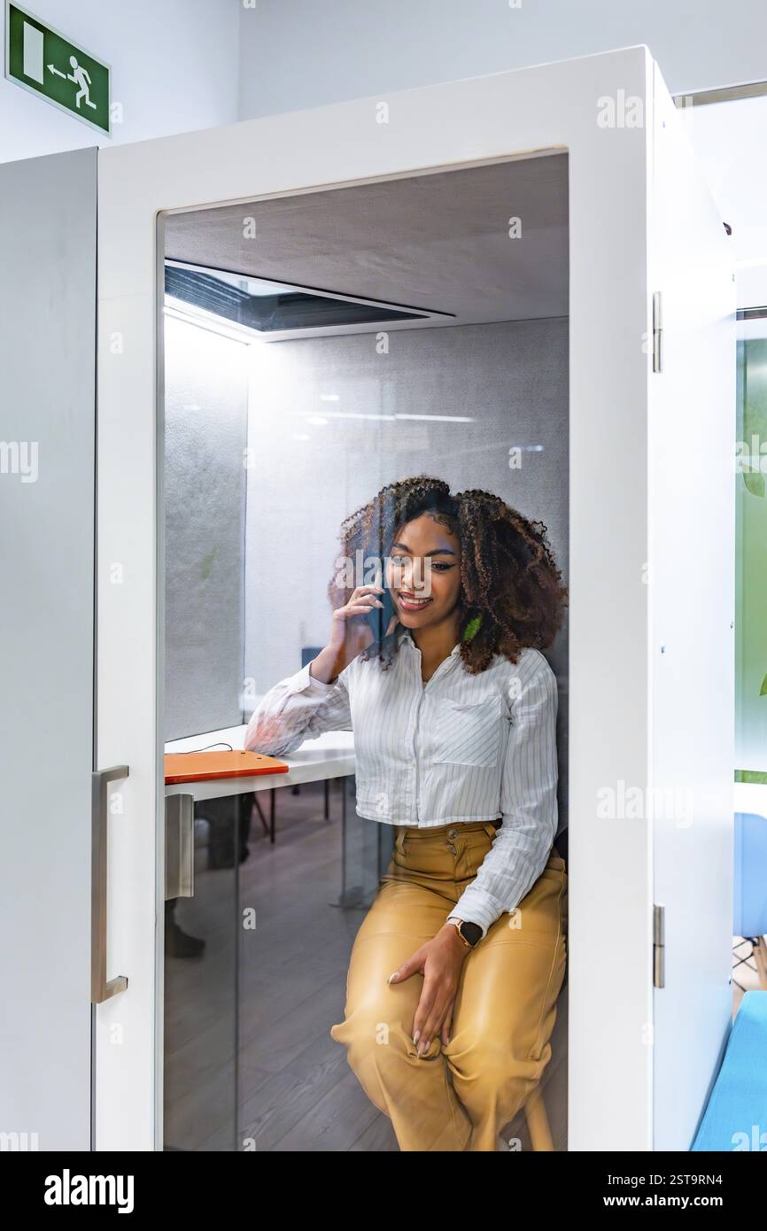 Jeune manager utilise son téléphone à l'intérieur d'une cabine dans un espace de coworking, profitant de l'intimité et du calme pour un travail concentré Banque D'Images
