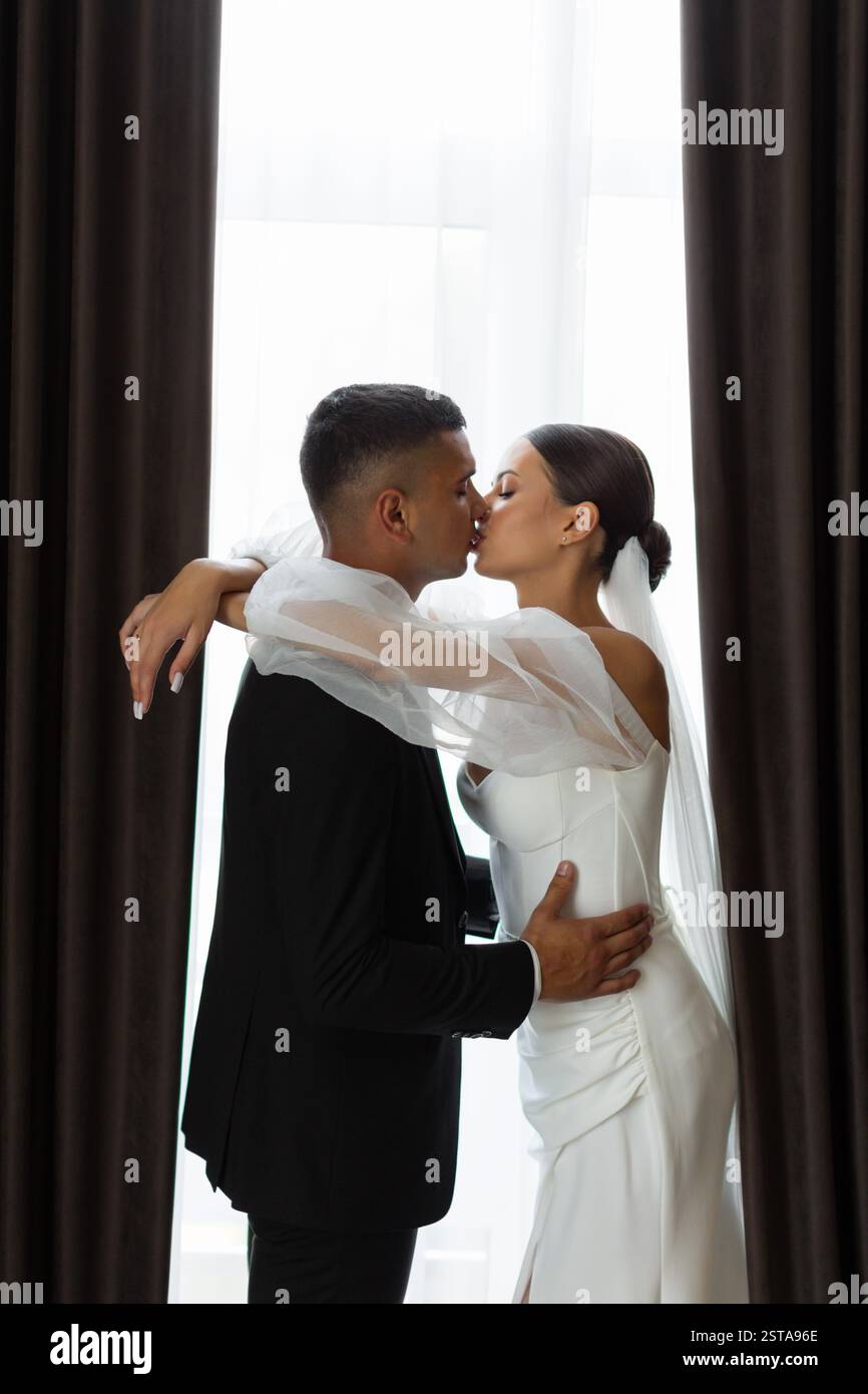 Portrait romantique de marié embrassant la mariée sur le front tout en embrassant près de la fenêtre le jour du mariage Banque D'Images
