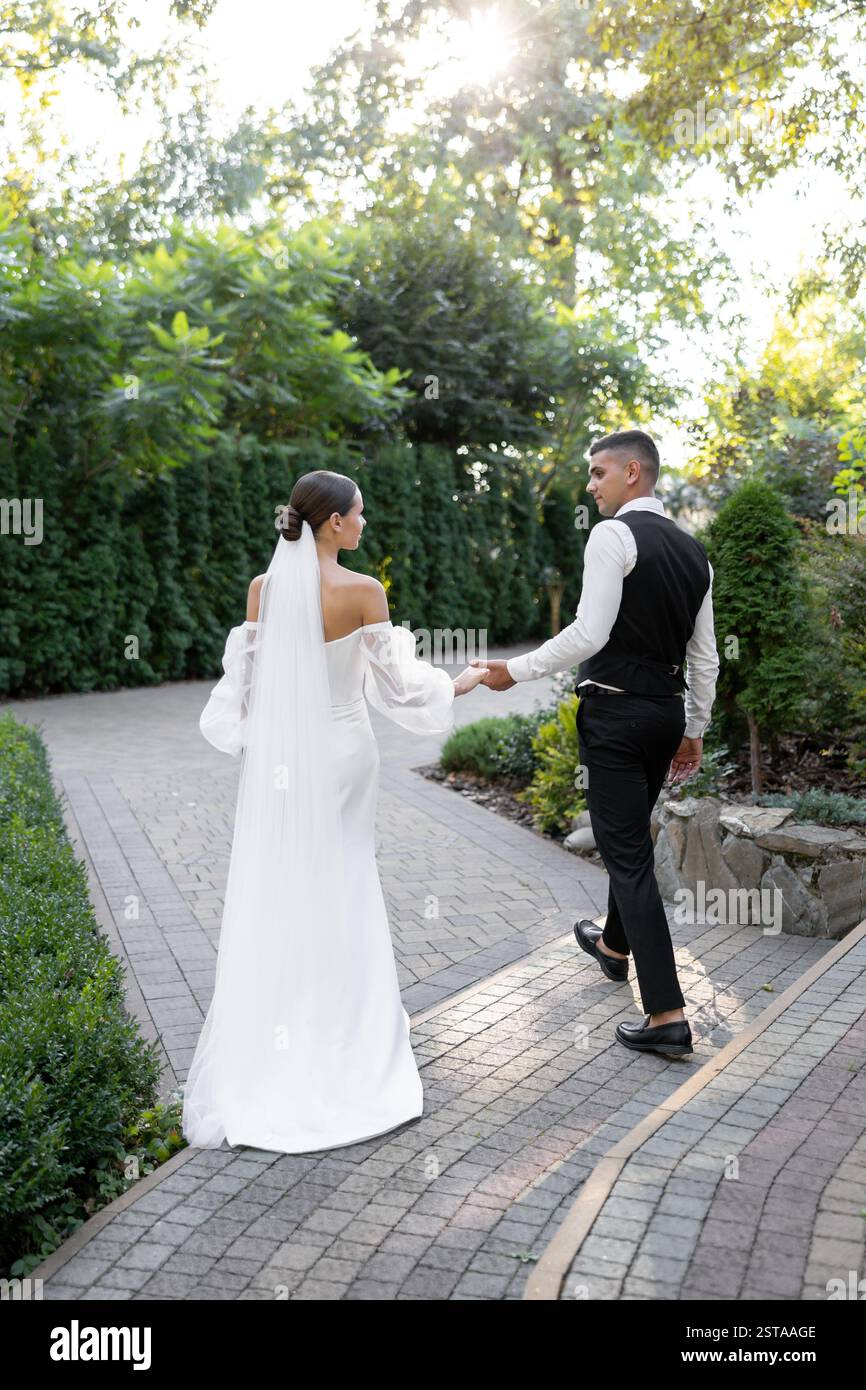 Couple de jeunes mariés se tenant la main et s'embrassant dans un parc, célébrant leur amour et leur engagement lors de leur cérémonie de mariage Banque D'Images Couple de jeunes mariés se tenant la main et s'embrassant dans un parc, célébrant leur amour et leur engagement lors de leur cérémonie de mariage Banque D'Images