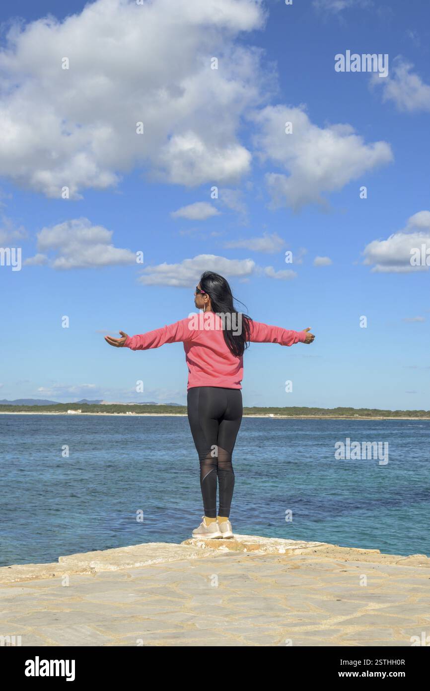 Une femme se tient avec ses bras ouverts grand, face à l'océan et profitant de la vue sereine, belle femme latina par la mer à Colonia San Jordi, Baleari Banque D'Images