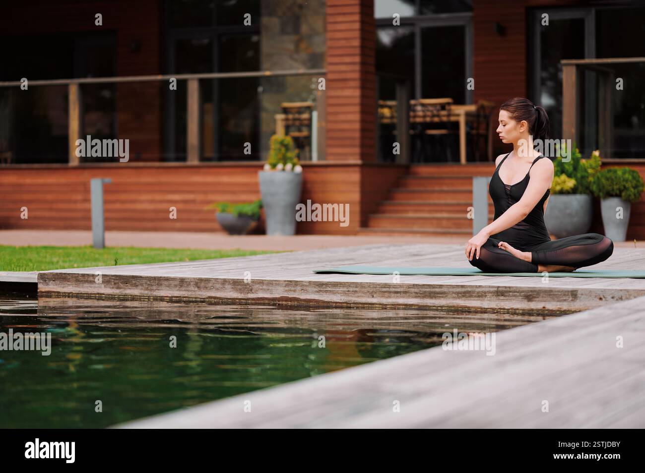 Jeune femme caucasienne effectue une torsion latérale assise dans la pose de lotus sur un tapis de yoga assis au bord de l'eau. Femme sportive pratiquant le yoga dans le parc. Femme doin Banque D'Images