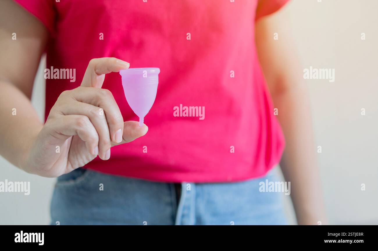 Femme portant une chemise rouge tenant une coupe menstruelle rose devant un fond blanc. Mise au point sélective. Banque D'Images