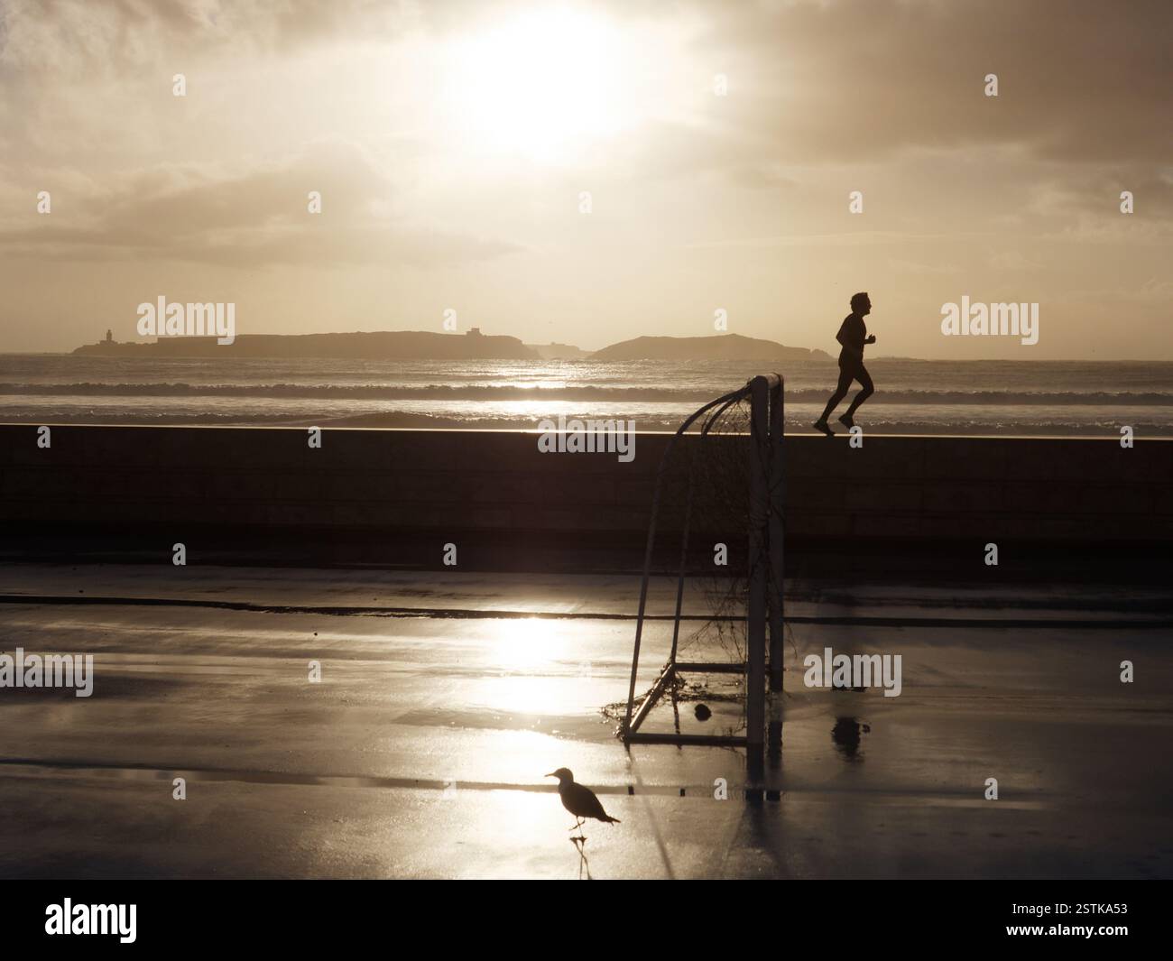 Jogger court sur le mur en silhouette après la pluie avec but de football au premier plan et mer et île derrière à Essaouira, Maroc. 18 février 2025 Banque D'Images