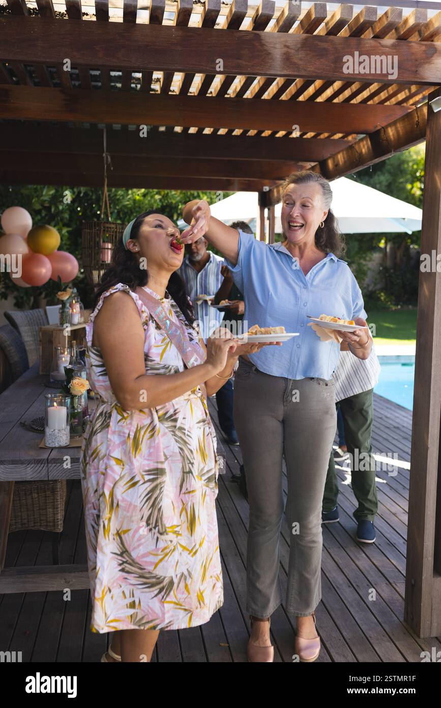 Senior diverse femme diverse amis seniors profitant de la fête en plein air, partageant de la nourriture et riant au bord de la piscine. Personnes âgées, amitié, célébration, rassemblement Banque D'Images