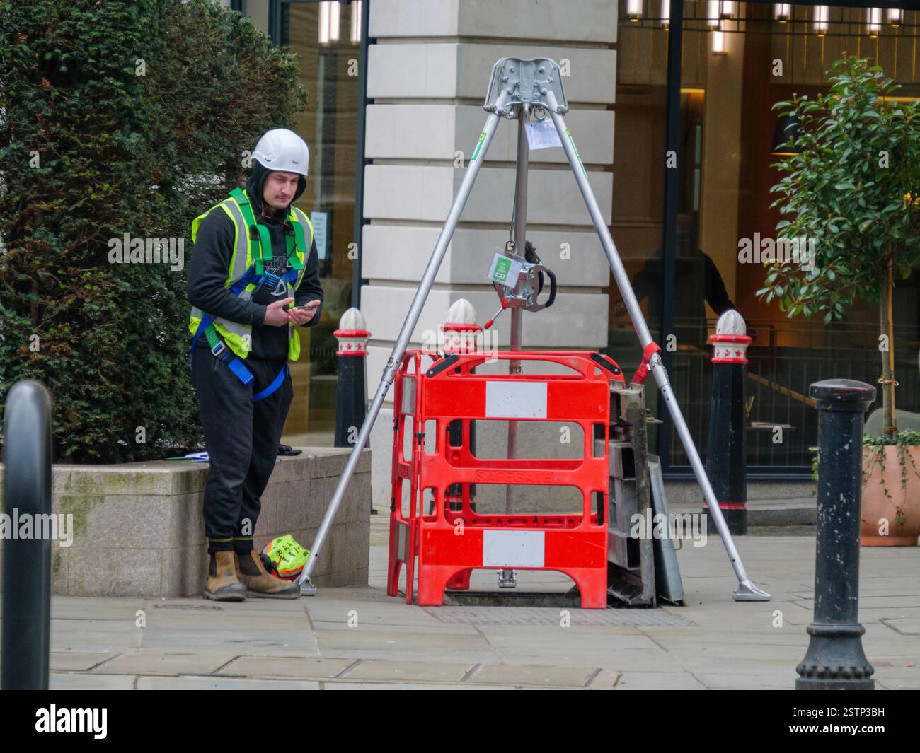 Travailleur portant un casque debout à côté de Manhole Cover soulevé avec des barrières équipement de sécurité et trépied de sauvetage en espace confiné avec treuil, centre de Londres Royaume-Uni Banque D'Images