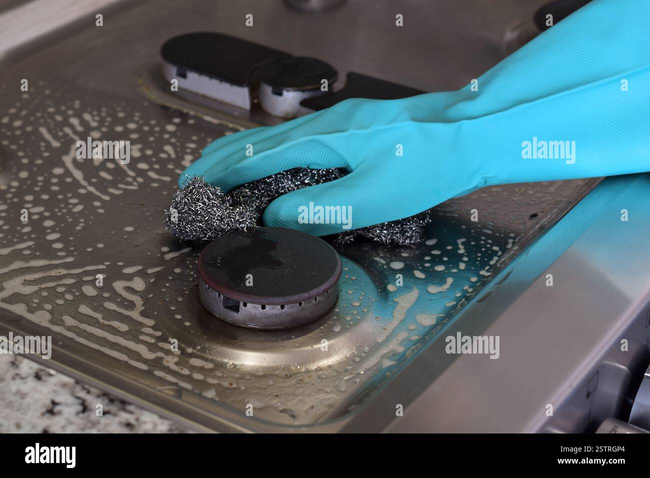 Main de la personne dans le gant en caoutchouc bleu nettoyant le dessus de la cuisinière à gaz avec épurateur de laine d'acier Banque D'Images