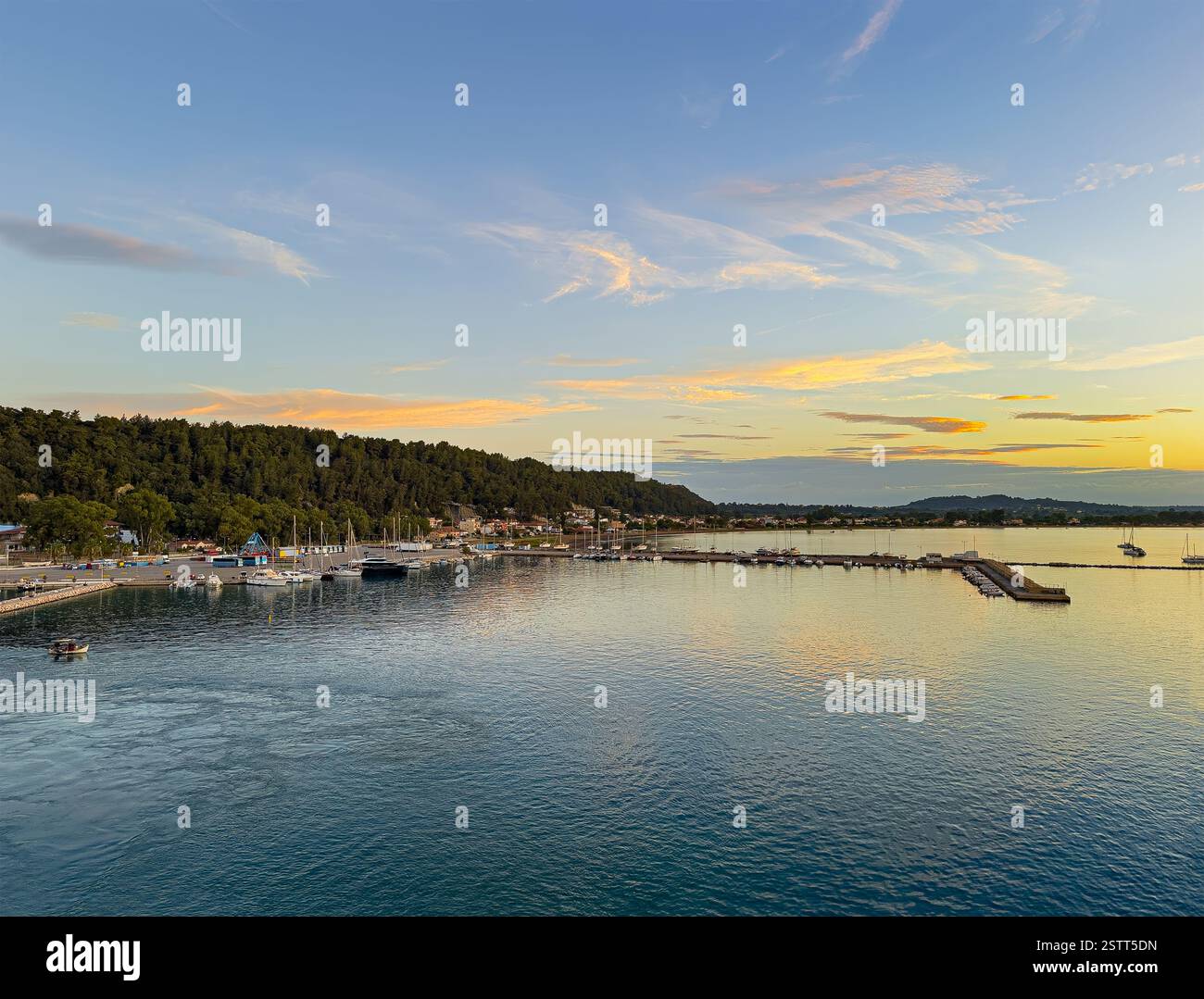 Katakolo, Grèce - 4 juillet 2024 : lever du soleil sur Katakolo Yacht Marina, Ilia, avec le rivage boisé derrière les bâtiments commerciaux le long de la promenade. Banque D'Images