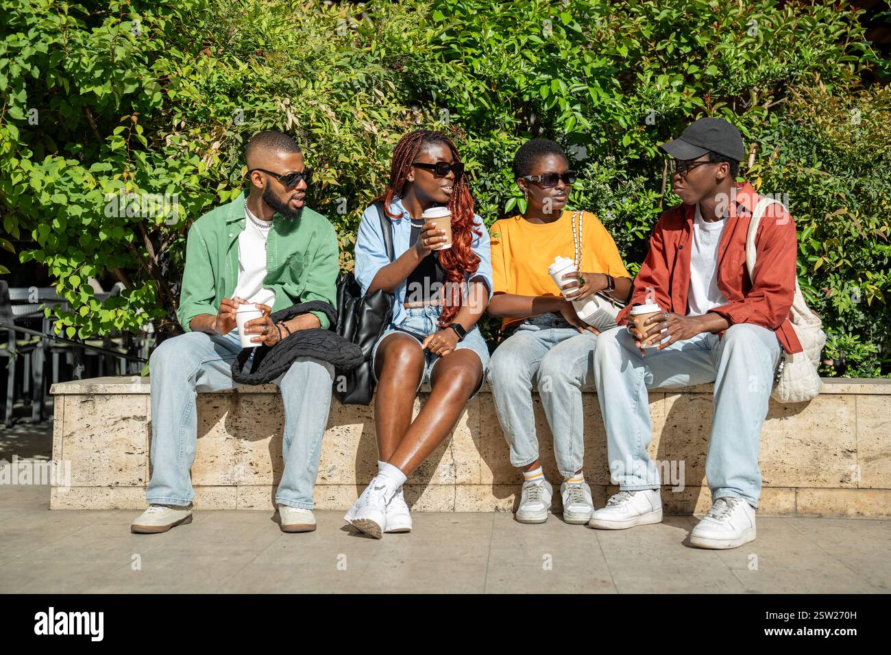 Groupe détendu d'amis afro-américains assis sur le banc du parc buvant du café tout en discutant Banque D'Images