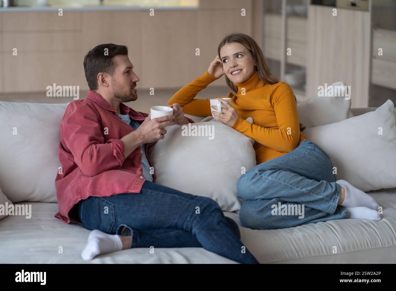 Un couple marié détendu se repose sur un canapé avec des tasses de café, discutant avec intérêt, planifiant l'avenir. Banque D'Images