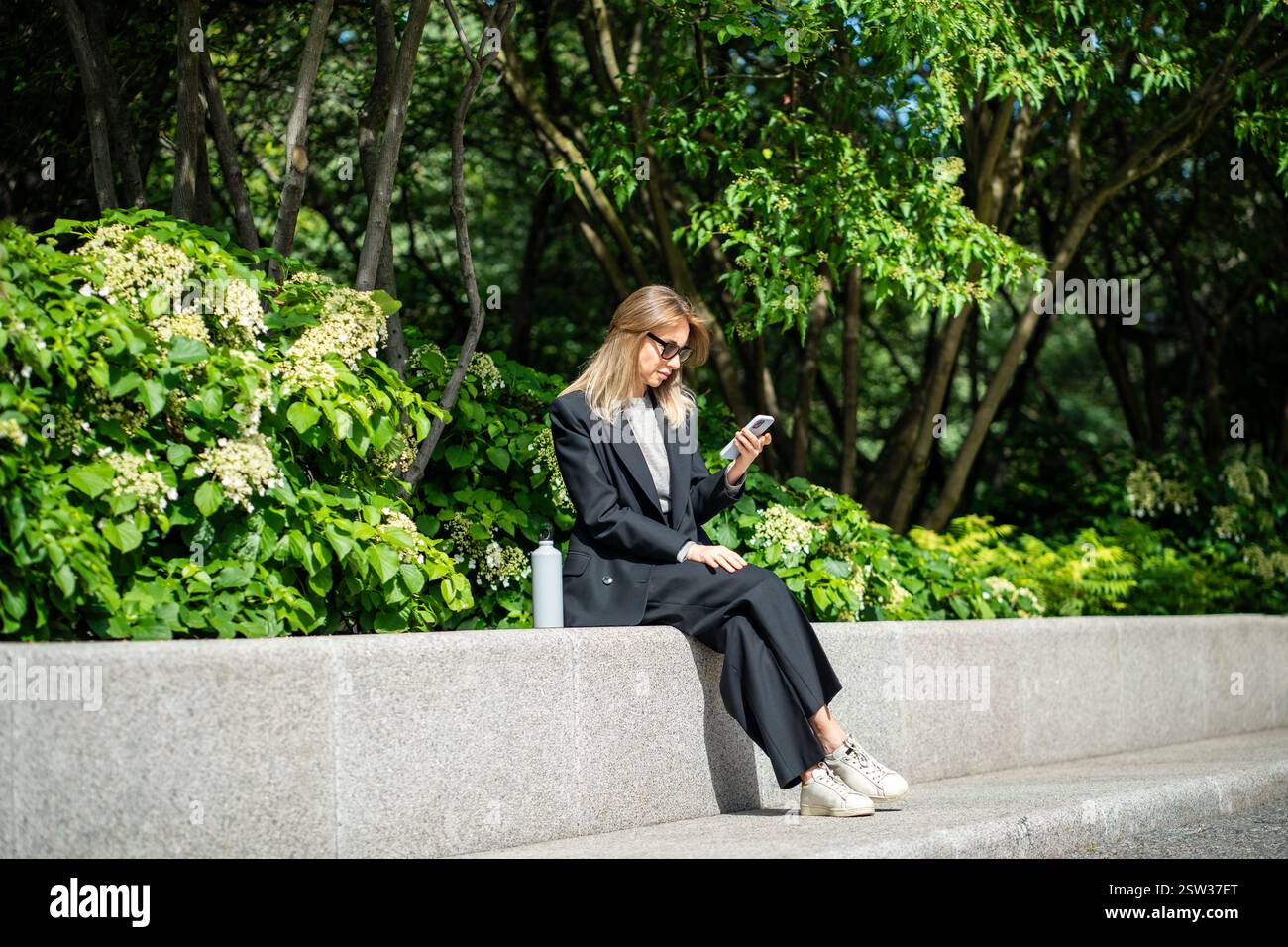 Femme d'affaires détendue dans des lunettes de soleil à l'aide d'un smartphone assis à l'extérieur près des plantes prenant la pause. Banque D'Images