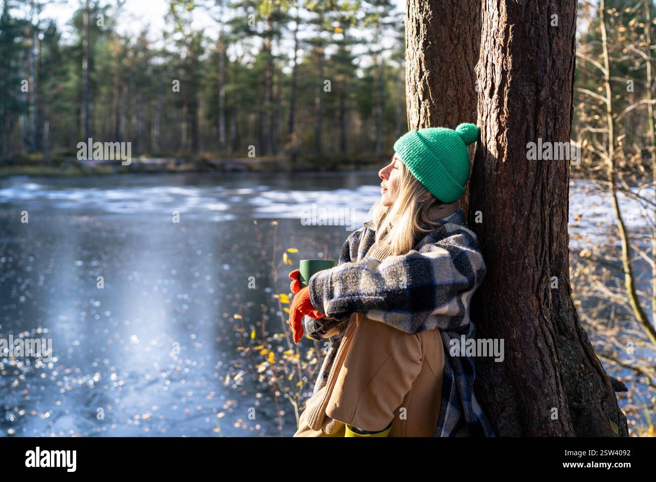 Femme calme assise près du tronc d'arbre dans la forêt d'hiver, regardant la rivière, profitant d'un passe-temps paisible à l'extérieur Banque D'Images