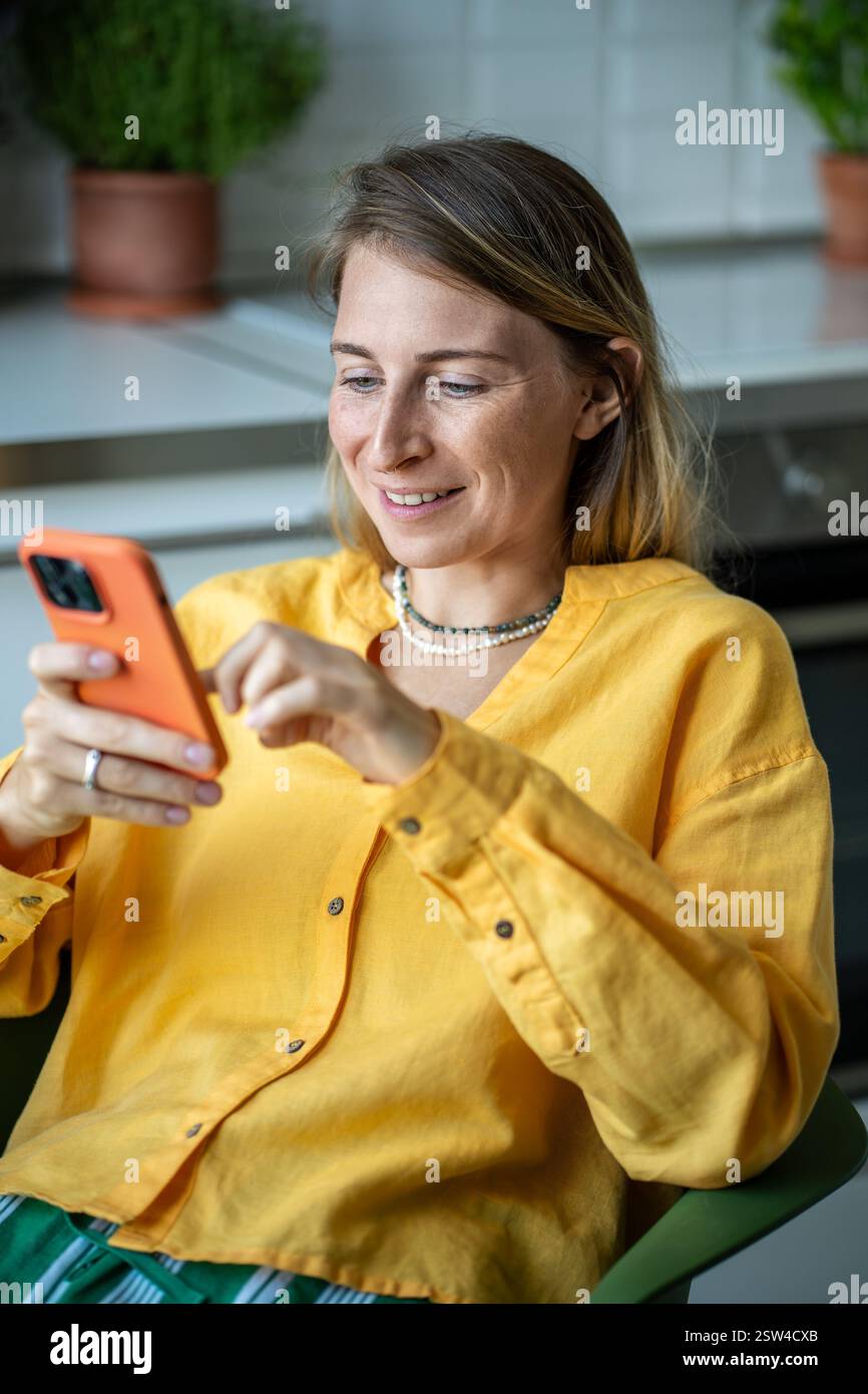Heureuse femme suédoise avec smartphone discutant avec des amis assis dans la chaise dans la cuisine et se relaxant. Banque D'Images