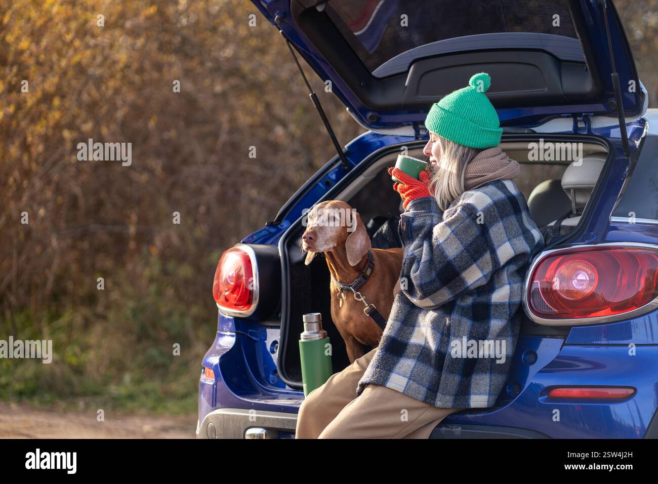 Femme propriétaire d'animal de compagnie buvant du thé assis dans le coffre de voiture avec chien appréciant la nature dans la forêt le week-end. Banque D'Images