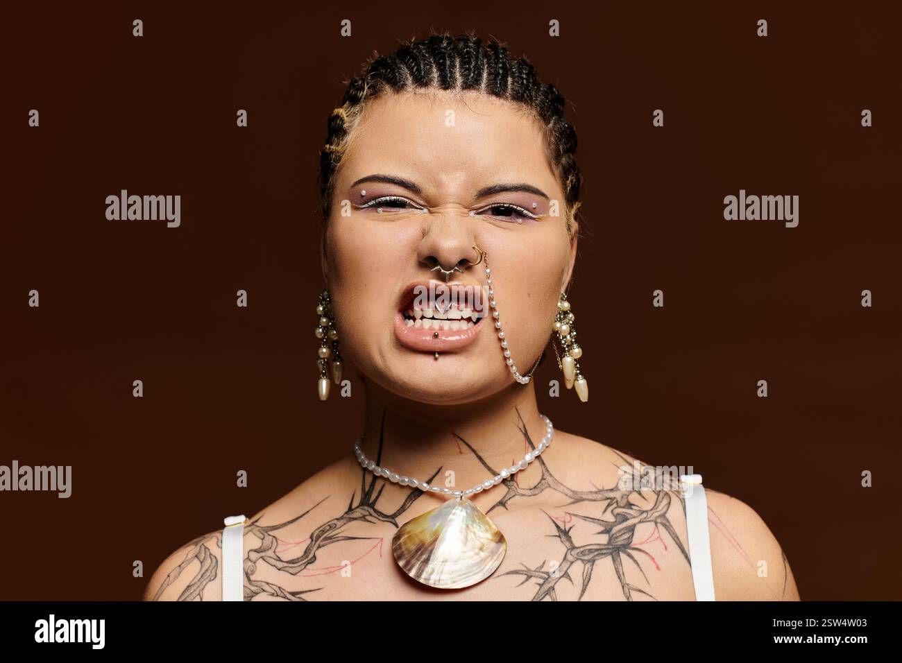 Une jeune femme affiche son style unique avec des tresses, des tatouages et des accessoires frappants dans un studio. Banque D'Images