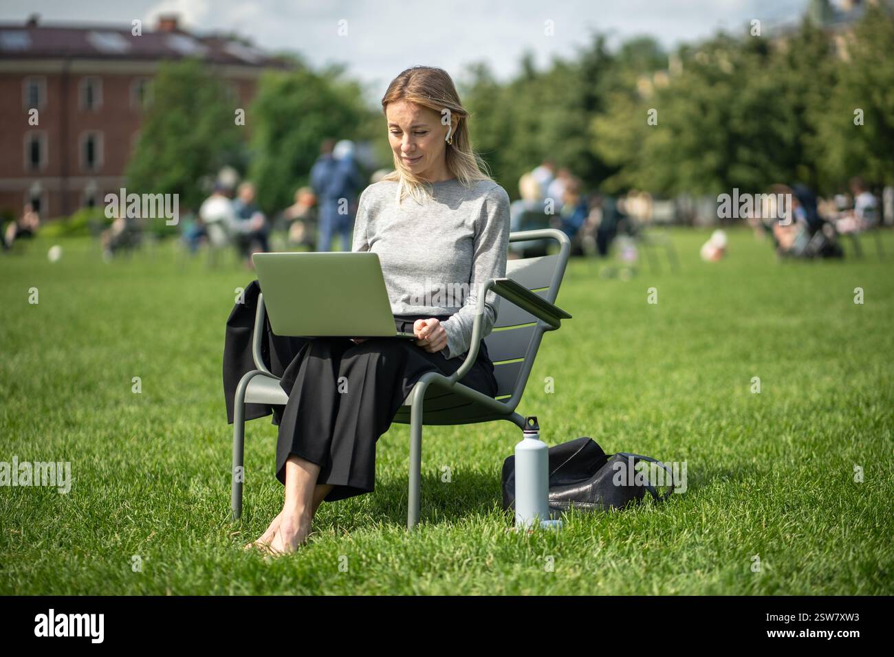 Femme souriante travaillant sur ordinateur portable avec des écouteurs sans fil, thermos dans le parc en été. Navigation sur Internet Banque D'Images