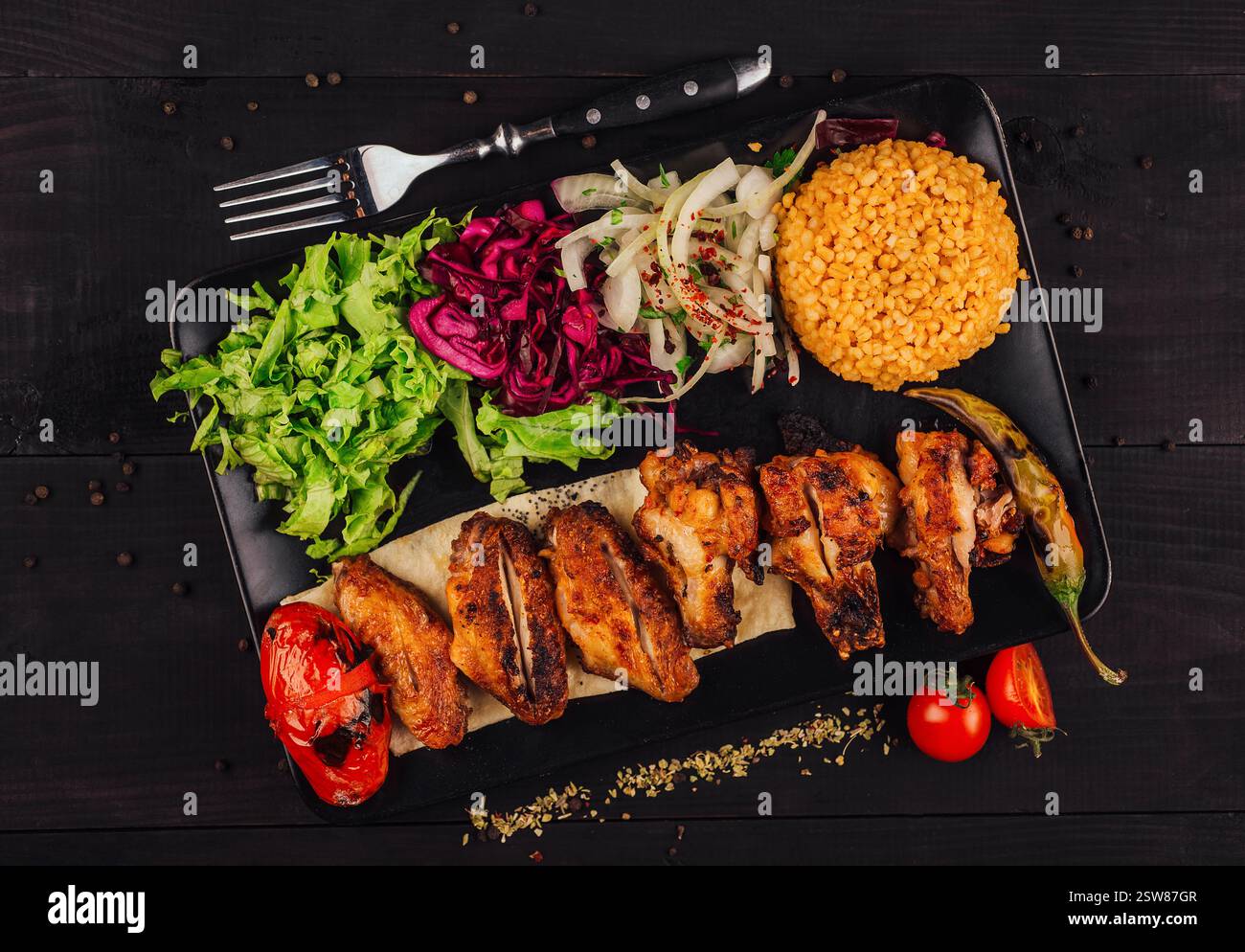 Ailes de poulet grillées servies sur assiette noire avec salade et bulgur sur fond de bois foncé Banque D'Images