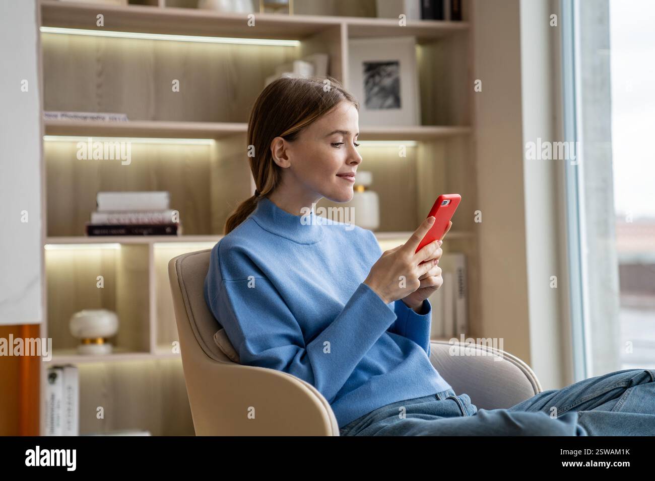 Femme souriante assise sur un fauteuil à la maison, en utilisant un téléphone portable, en lisant, en écrivant un message, en discutant. Banque D'Images