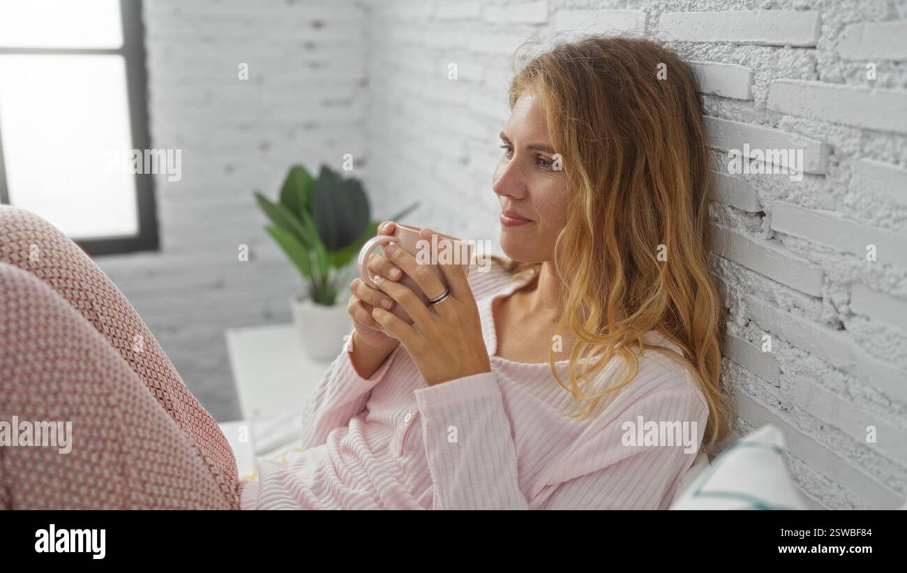 Jeune femme blonde profite d'une tasse de café tout en se relaxant dans sa chambre aux murs de briques blanches, vêtue d'un pyjama rose. Banque D'Images