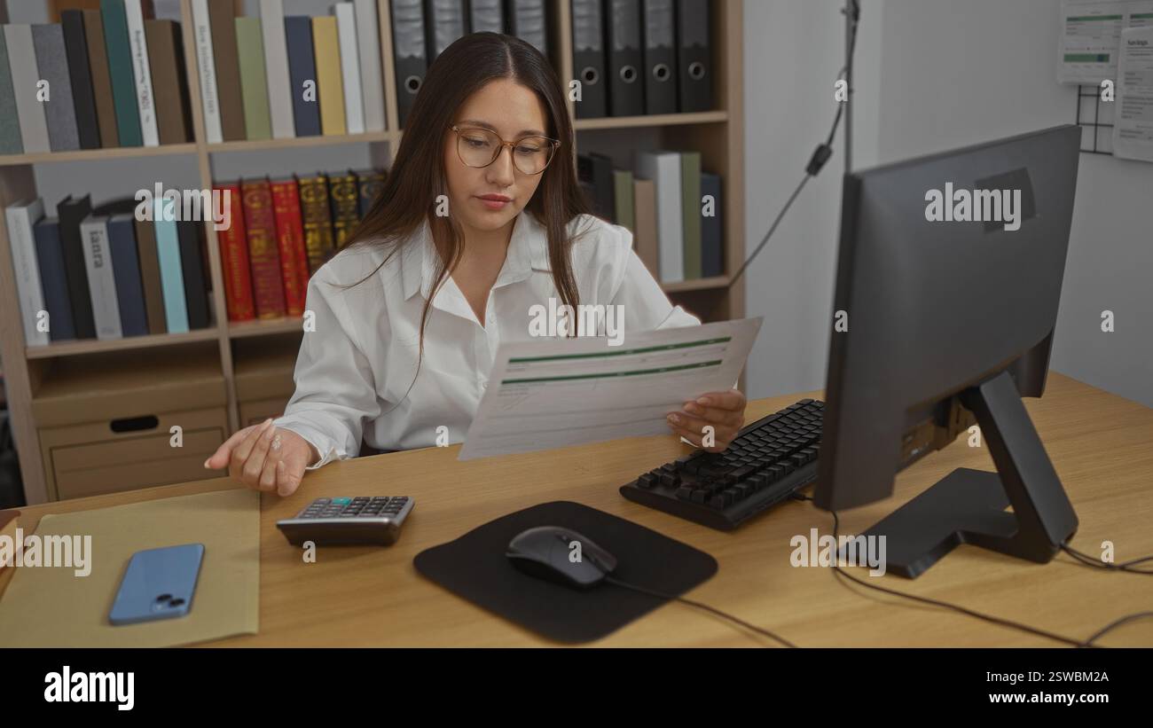 Jeune femme travaillant à un bureau dans une pièce de bureau avec des papiers, un ordinateur et des étagères pleines de livres et de classeurs en arrière-plan Banque D'Images