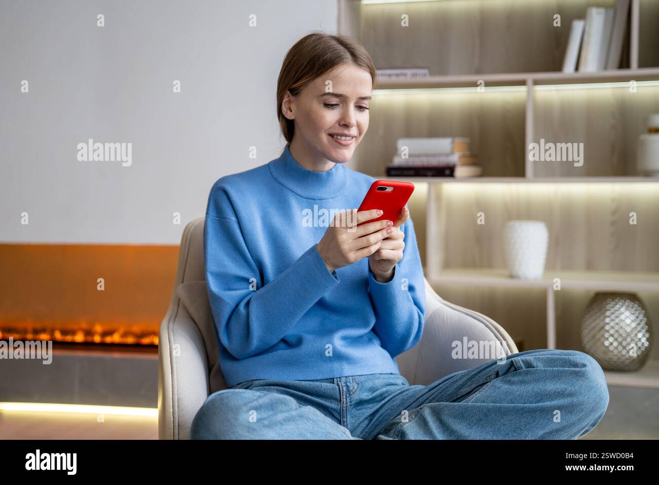 Femme souriante assise sur un fauteuil à la maison, en utilisant un téléphone portable, en lisant, en écrivant un message, en discutant. Banque D'Images