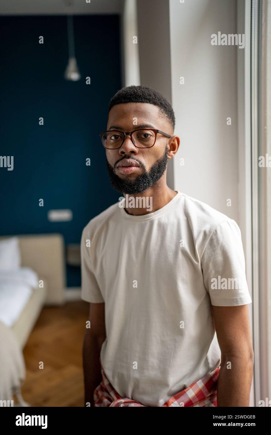 Portrait homme barbu afro-américain sérieux en lunettes dans un t-shirt blanc près de la fenêtre à la maison Banque D'Images