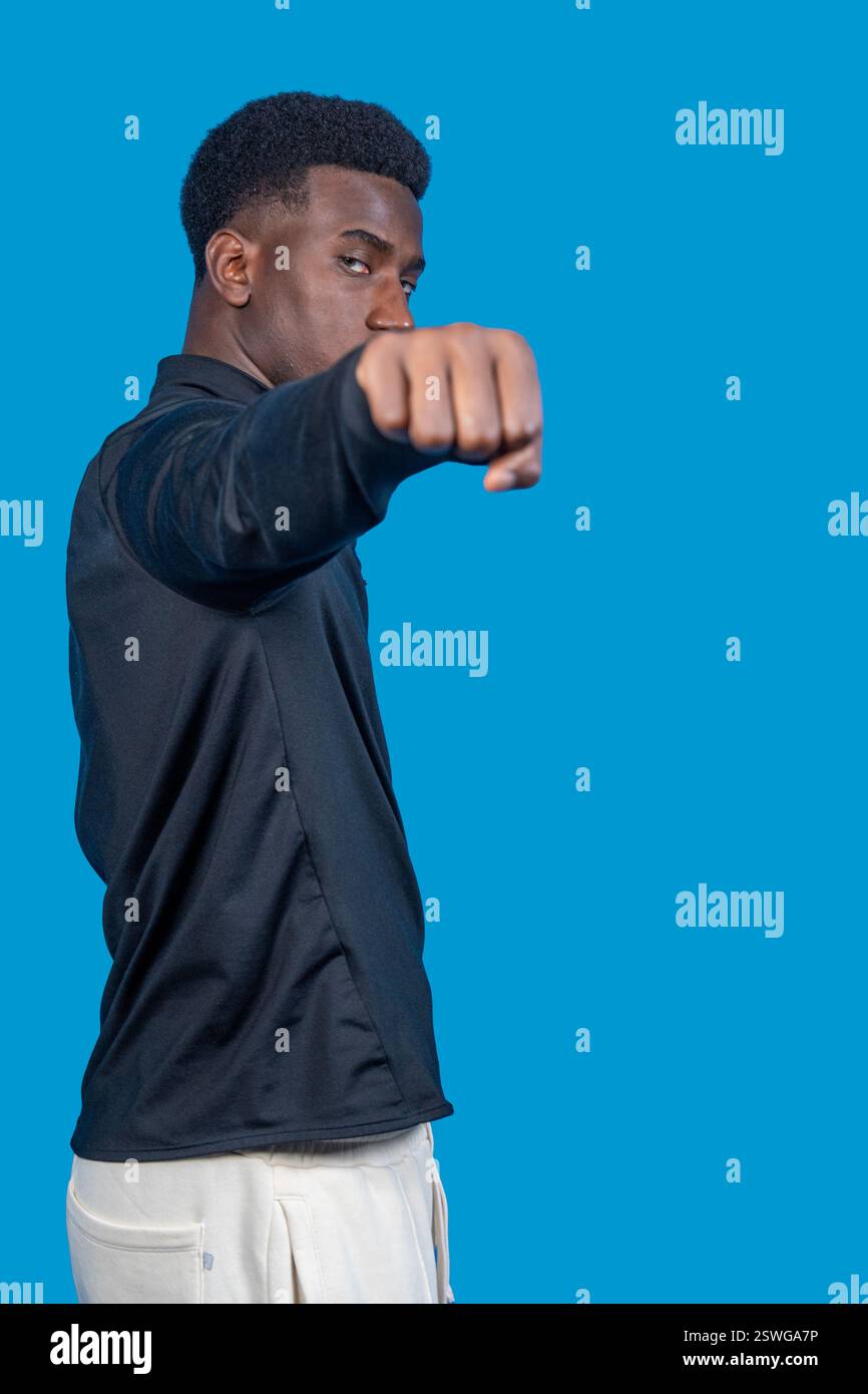 Jeune athlète sérieux pratiquant la boxe jetant un coup de poing sur fond bleu Banque D'Images