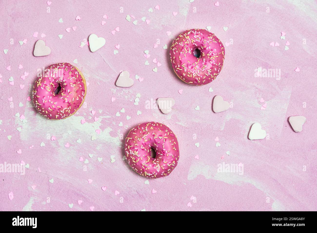 Beignets, Berliner, Krapfen avec glaçage au sucre rose sur fond texturé rose, décoré de sucre rose et blanc et de coeurs en papier, vue de dessus Banque D'Images