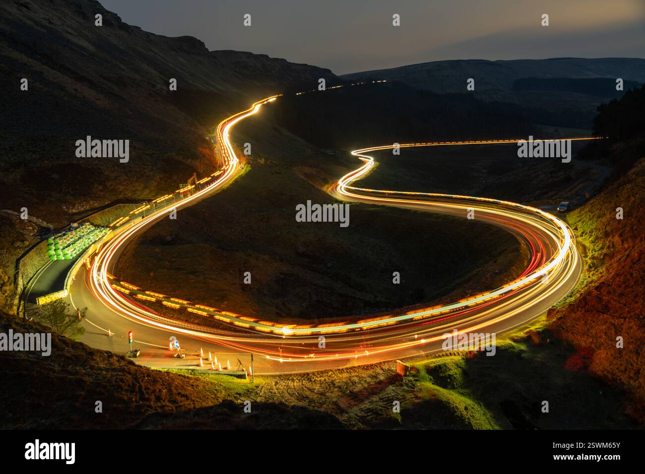 Une photographie longue exposition d'une route de montagne sinueuse la nuit, avec des traînées de lumière de voiture créant un chemin lumineux à travers le paysage. La scène est surr Banque D'Images