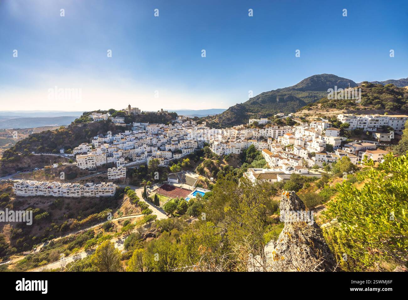 Une vue imprenable sur un charmant village blanc accroché à une colline, entouré de verdure luxuriante et de montagnes majestueuses. La lumière du soleil brillante met en évidence Banque D'Images