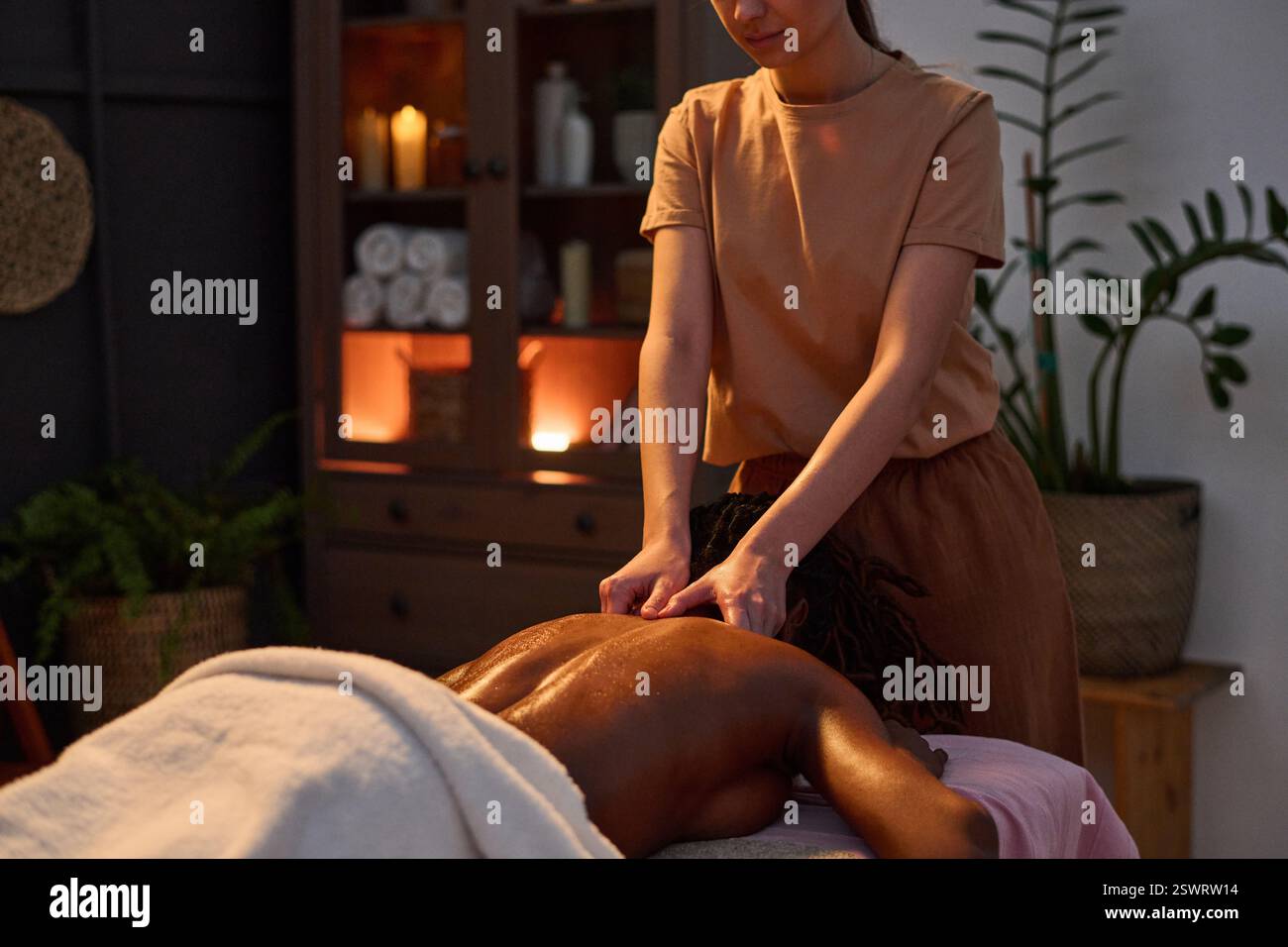 Grand plan de masseuse méconnaissable pétrissant le dos de la femme à l'aide d'huile chauffante, femme afro-américaine allongée sur le ventre et profitant d'un massage relaxant dans une pièce faiblement éclairée Banque D'Images