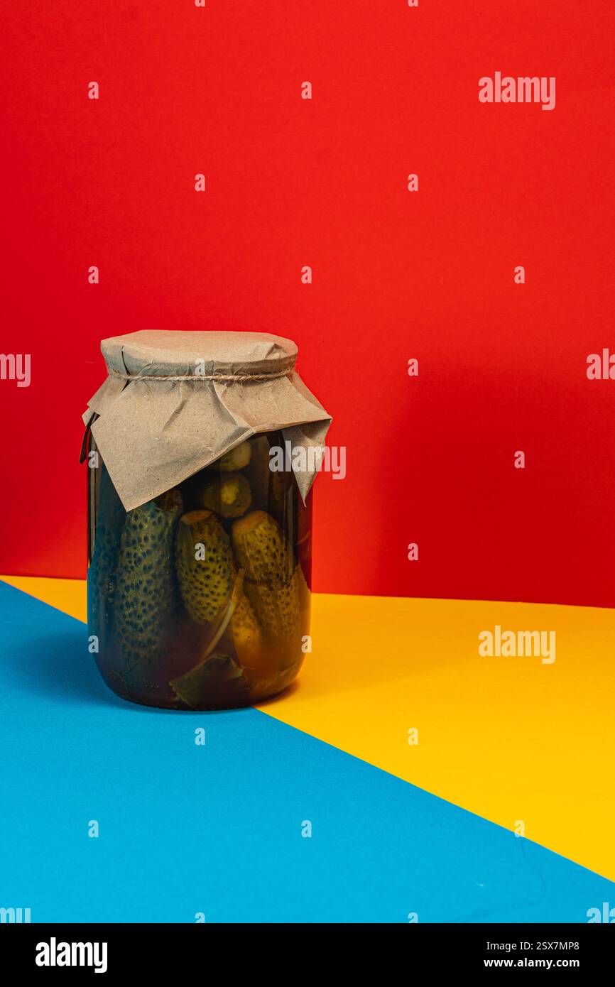 Un pot de concombres marinés maison est présenté sur un fond rouge et bleu saisissant, soulignant le contraste des légumes conservés et Banque D'Images