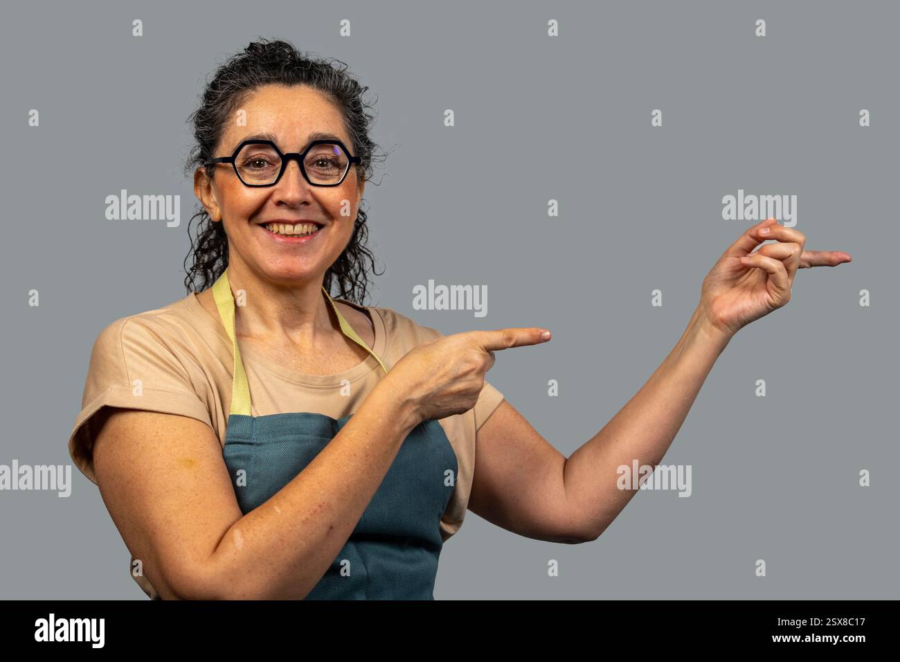 Femme mature heureuse portant un tablier et des lunettes pointant de côté avec des index et montrant quelque chose sur fond gris Banque D'Images