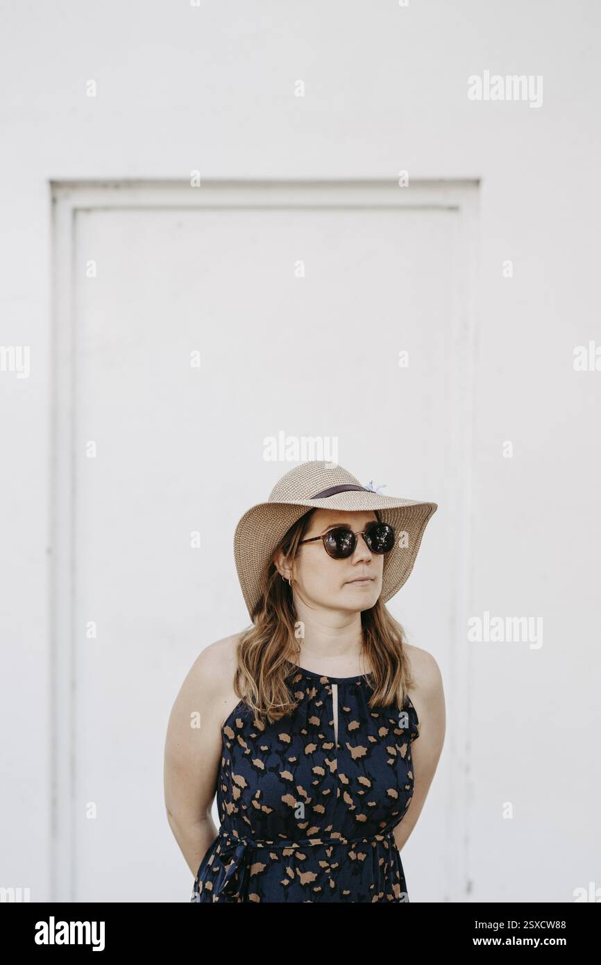 Femme, 30 ans et plus, avec un chapeau de soleil et des lunettes de soleil devant un mur blanc Banque D'Images