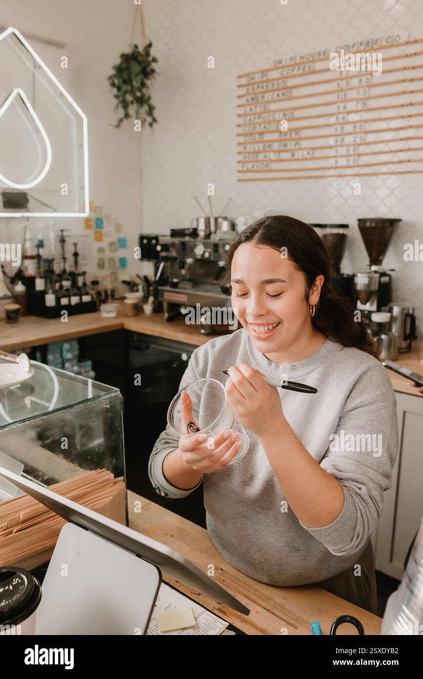 Barista souriant écrivant sur une tasse tout en prenant une commande au comptoir du café Banque D'Images
