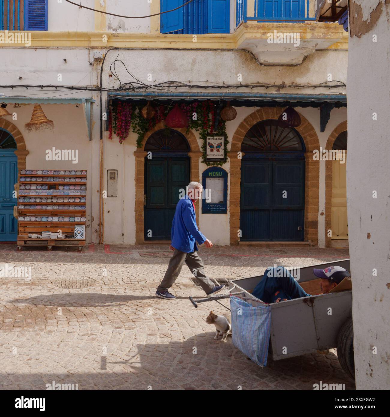 Un vieil homme marche dans une rue aux portes bleues tandis qu'un autre est assis dans un chariot à bagages observant les gens, à Essaouira, Maroc, février 2025 Banque D'Images