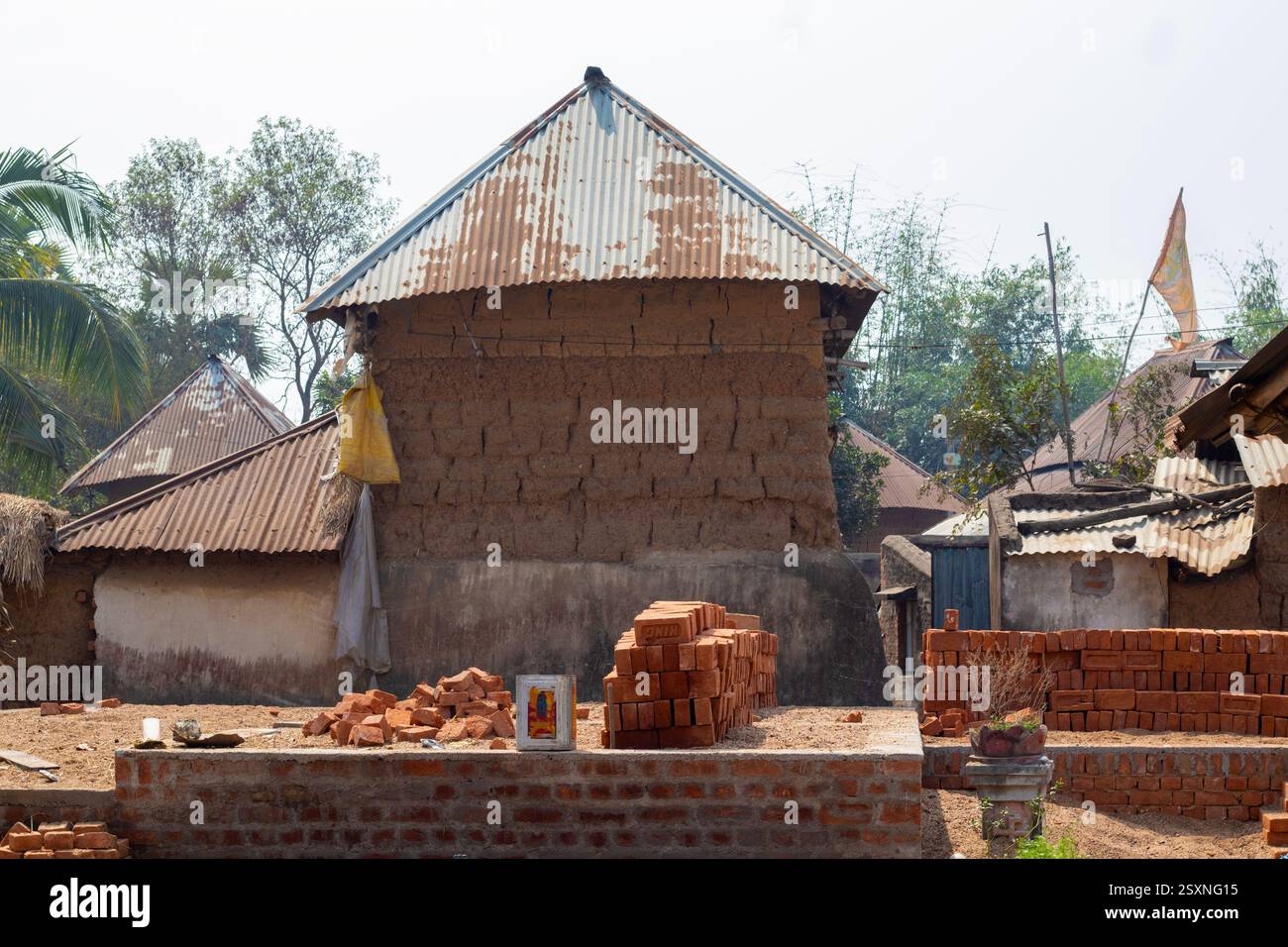 Une maison de boue traditionnelle de village indien avec un toit en métal ondulé, entourée d'arbres, et des structures rurales Banque D'Images