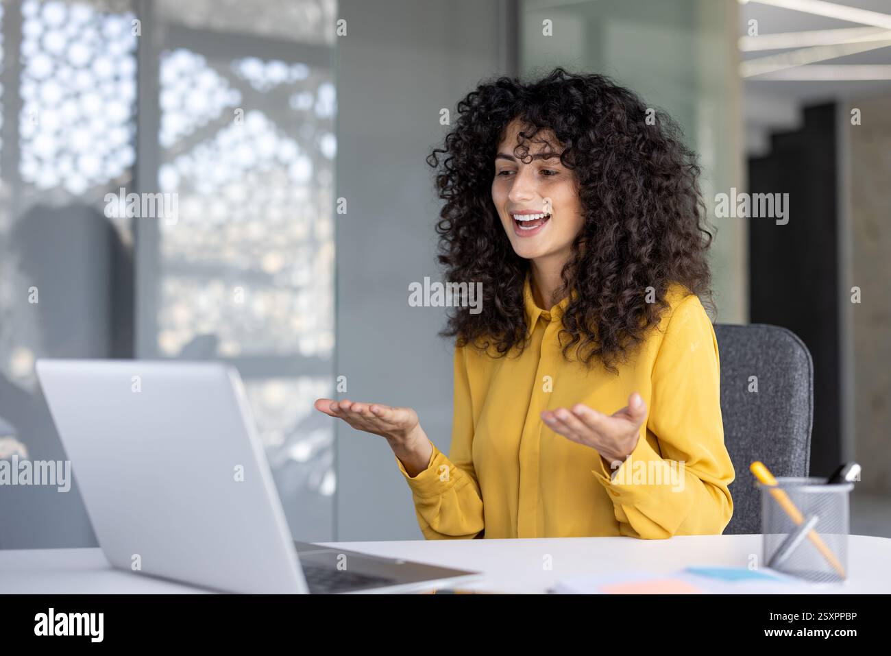 Une femme sourit tout en faisant des gestes pendant un appel vidéo dynamique sur son ordinateur portable dans un environnement de bureau moderne Banque D'Images