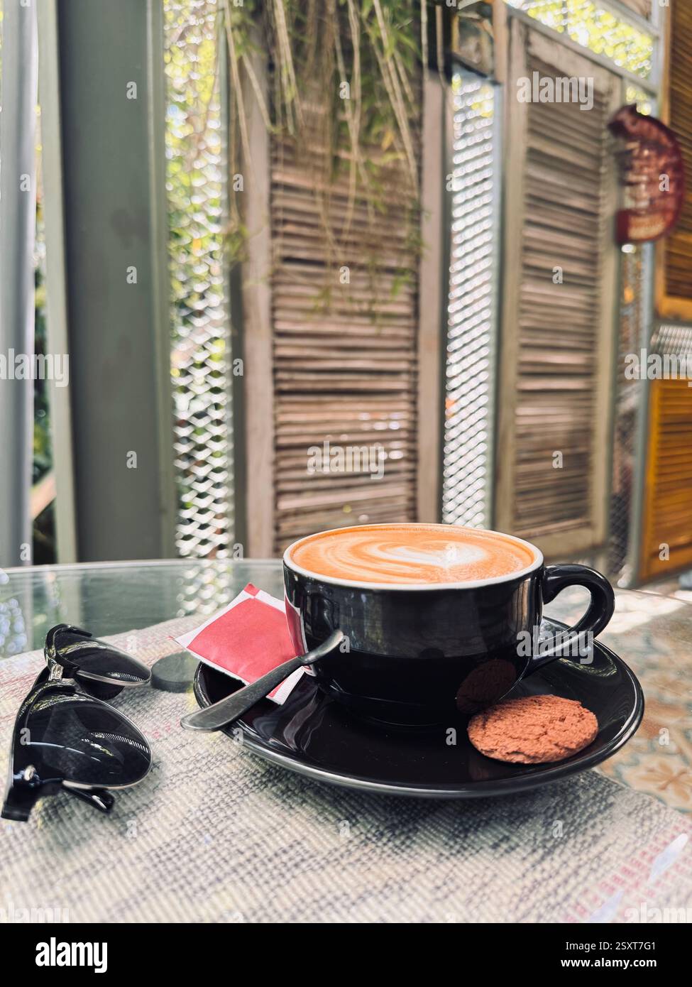Une scène de café confortable avec une tasse de café avec latte art, un biscuit sur le côté, et des lunettes de soleil reposant sur une table. L'arrière-plan comprend le te en bois - Image de stock capturée avec un smartphone
