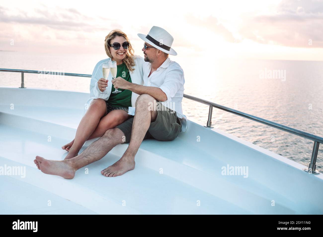 Un jeune couple marié rencontre le coucher du soleil à la poupe du navire. Une femme souriante en lunettes de soleil prit des verres de champagne avec son mari. Un homme an Banque D'Images