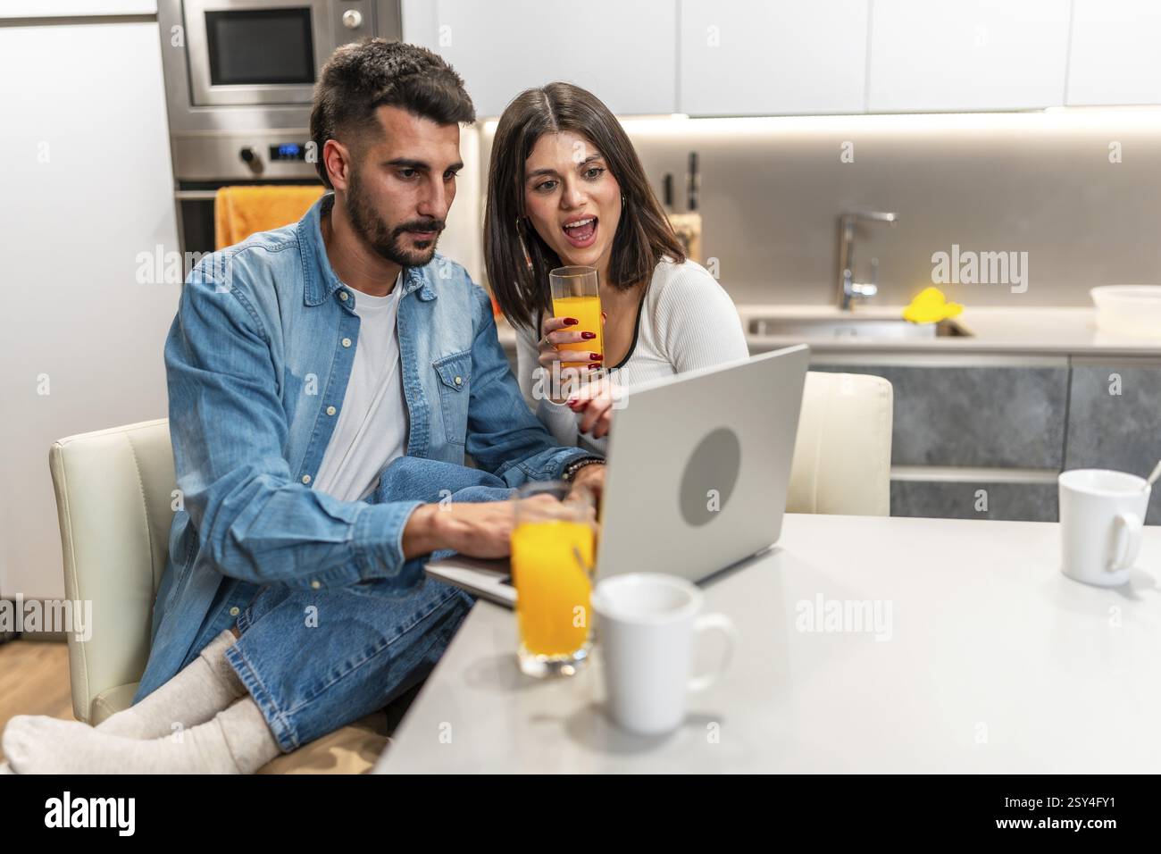 Jeune couple utilisant un ordinateur portable et buvant du jus d'orange dans une cuisine moderne, profitant du temps ensemble à la maison Banque D'Images