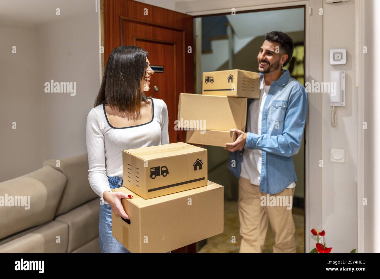 Jeune couple portant des boîtes en carton tout en entrant dans leur nouvelle maison, en souriant et en se regardant Banque D'Images