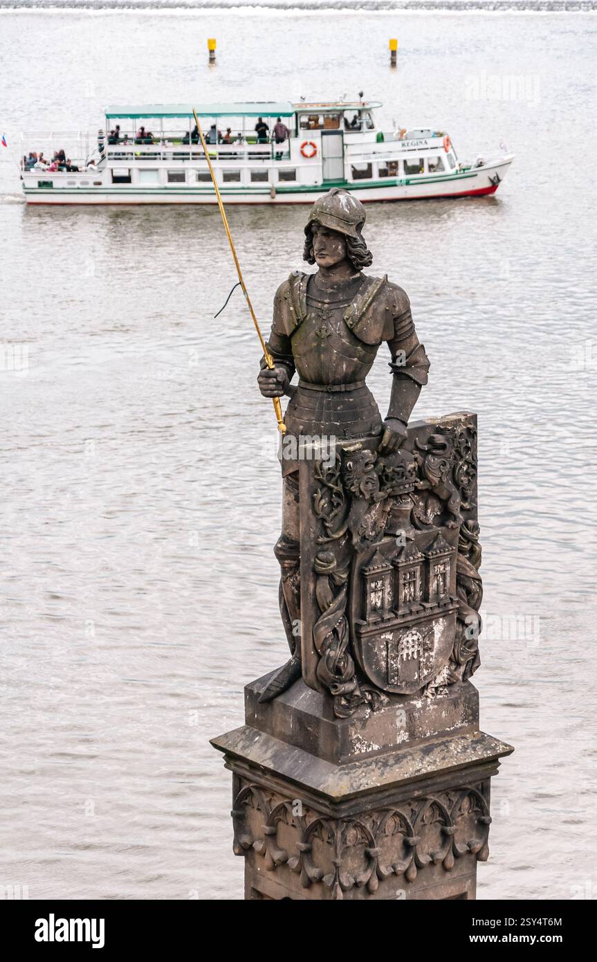 Prague, République tchèque - 12 juin 2016 : une statue d'un chevalier tenant une lance se dresse bien en vue près de la rivière Vltava, avec un bateau d'excursion passant dans le b Banque D'Images